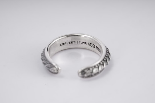 A silver open-ended ring with intricate patterns on the outer surface. The inside is engraved with the text 'COPPERTIST.WU S925 ANT' indicating sterling silver. The design is elegant and detailed, complemented by a smooth inner finish.