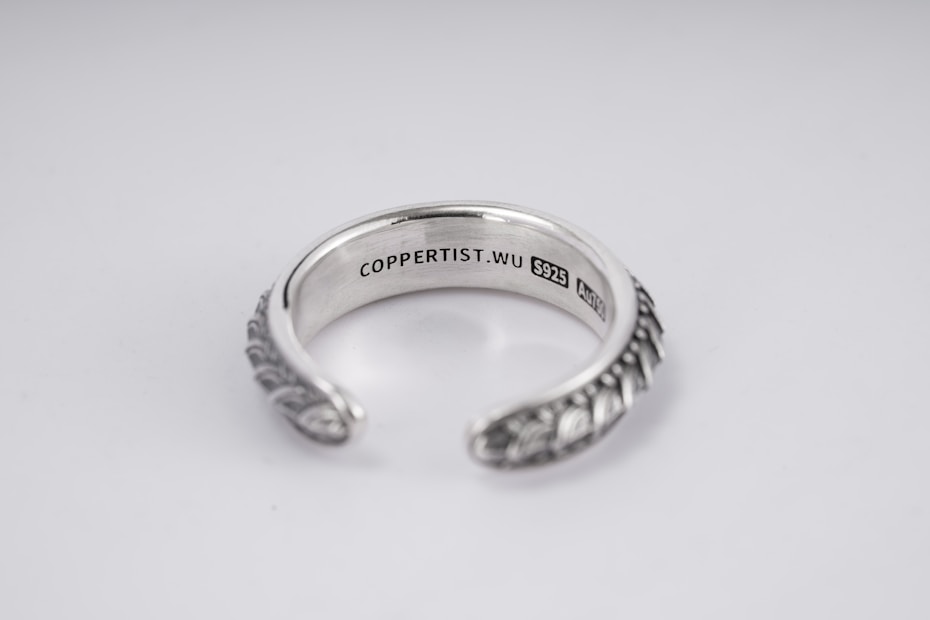 A silver open-ended ring with intricate patterns on the outer surface. The inside is engraved with the text 'COPPERTIST.WU S925 ANT' indicating sterling silver. The design is elegant and detailed, complemented by a smooth inner finish.