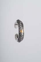 An intricately designed silver ring featuring a textured scale-like pattern and a central gold accent. The ring appears suspended against a plain, light grey background, highlighting its ornate craftsmanship and metallic sheen.