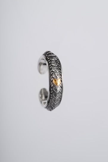 An intricately designed silver ring featuring a textured scale-like pattern and a central gold accent. The ring appears suspended against a plain, light grey background, highlighting its ornate craftsmanship and metallic sheen.