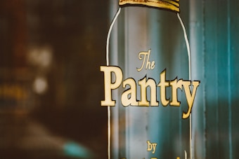 A large glass jar with the words 'The Pantry' written on it in yellow letters. The jar is slightly transparent and has a rustic, vintage aesthetic. The background appears to be a blurred, indoor setting.