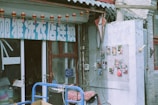 A storefront with a sign in a foreign script above the entrance, adorned with small red lanterns hanging from strings. To the right, a wall displays various colorful photographs arranged in a grid. The building facade features weathered bricks and a decorative grey roof with circular tiles. Some cables and a box cart with items are visible in the foreground.