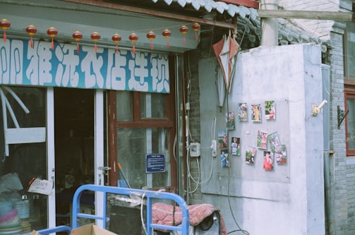 A storefront with a sign in a foreign script above the entrance, adorned with small red lanterns hanging from strings. To the right, a wall displays various colorful photographs arranged in a grid. The building facade features weathered bricks and a decorative grey roof with circular tiles. Some cables and a box cart with items are visible in the foreground.