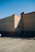A parking lot with marked spaces adjacent to a large beige building. The building casts a long shadow on the ground. The sky is clear and blue. A lamp post is visible near the building wall, and a small part of a car is partly visible to the left of the image.