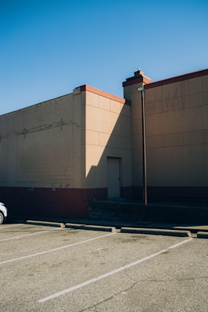 A parking lot with marked spaces adjacent to a large beige building. The building casts a long shadow on the ground. The sky is clear and blue. A lamp post is visible near the building wall, and a small part of a car is partly visible to the left of the image.