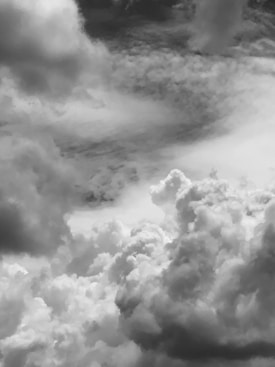 A dramatic view of large, fluffy clouds filling the sky. The clouds are layered, with some appearing wispy and others dense and towering. The image is in black and white, emphasizing the texture and contrast of the cloud formations.