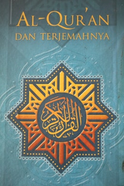 The cover features a geometric star design with intricate patterns in orange and gold, centered on a blue background. Arabic calligraphy is prominently displayed in the center within the geometric pattern. Above the design, the title 'Al-Qur'an Dan Terjemahnya' is printed in yellow text.