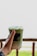 A hand holding a plastic cup with a green beverage, topped with foam and an opaque lid. The cup has a label that reads '38.1 Slow Bar.' The background is a blurred outdoor setting with some wooden railing and greenery.