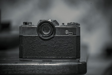 A vintage film camera sits prominently on a wooden surface, displaying its textured body and classic design with visible dials and lens. The monochromatic tone adds a nostalgic feel, enhancing the old-fashioned aesthetic.