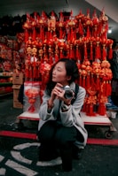 A person crouches while holding a camera amidst a vibrant display of red and gold Chinese New Year decorations. Behind them are an array of traditional hanging ornaments, featuring intricate designs and tassels, creating a festive atmosphere.