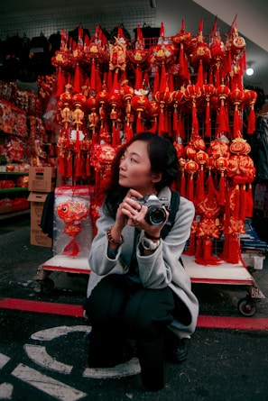 A person crouches while holding a camera amidst a vibrant display of red and gold Chinese New Year decorations. Behind them are an array of traditional hanging ornaments, featuring intricate designs and tassels, creating a festive atmosphere.