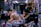 A tattoo artist is concentrated on completing a large, intricate tattoo on a client's back in a busy tattoo studio. Various equipment, supplies, and tattoo designs adorn the workspace, while the walls are filled with art and posters.