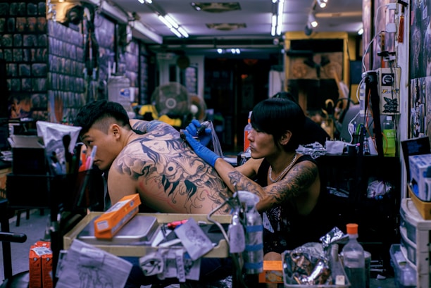 A tattoo artist is concentrated on completing a large, intricate tattoo on a client's back in a busy tattoo studio. Various equipment, supplies, and tattoo designs adorn the workspace, while the walls are filled with art and posters.