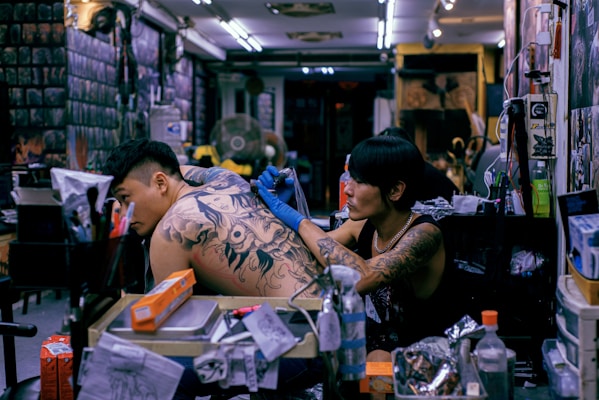 A tattoo artist is concentrated on completing a large, intricate tattoo on a client's back in a busy tattoo studio. Various equipment, supplies, and tattoo designs adorn the workspace, while the walls are filled with art and posters.