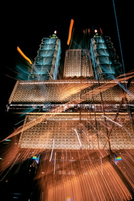 A dynamic image featuring two tall, illuminated towers with intricate, glowing architectural details against a dark sky. The light trails and sparks add a vibrant, energetic feel to the scene, creating a sense of movement and excitement.