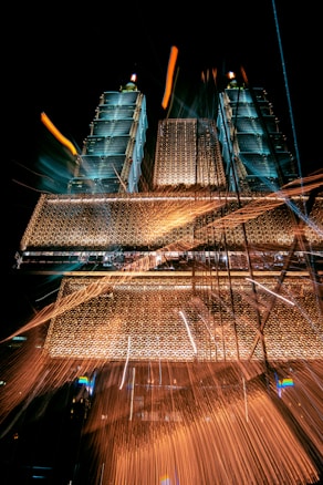 A dynamic image featuring two tall, illuminated towers with intricate, glowing architectural details against a dark sky. The light trails and sparks add a vibrant, energetic feel to the scene, creating a sense of movement and excitement.