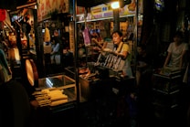 A bustling street food market scene with a vendor handling corn cobs on skewers. The vendor is in an apron, engaging with customers, while surrounding stalls display colorful signs in a foreign script. The warm glow of the market lights creates a vibrant atmosphere, with several people visible in the dimly lit background.