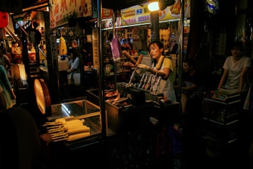 A bustling street food market scene with a vendor handling corn cobs on skewers. The vendor is in an apron, engaging with customers, while surrounding stalls display colorful signs in a foreign script. The warm glow of the market lights creates a vibrant atmosphere, with several people visible in the dimly lit background.