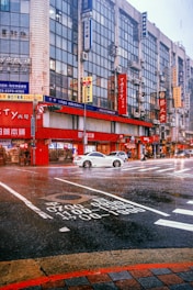 A busy urban street corner features tall buildings with numerous signs in various languages, primarily Asian script. A white car is stopped in the middle of an intersection with visible road markings. The scene takes place during rainfall, adding a wet sheen to the street. Pedestrians and possibly a cyclist or motorcyclist are also present.