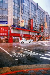 A busy urban street corner features tall buildings with numerous signs in various languages, primarily Asian script. A white car is stopped in the middle of an intersection with visible road markings. The scene takes place during rainfall, adding a wet sheen to the street. Pedestrians and possibly a cyclist or motorcyclist are also present.