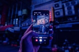 A hand holds a smartphone displaying a vibrant street scene captured in low light. Neon signs with apparent Japanese characters are visible in the photograph on the phone screen, while the background is dark and filled with indistinct urban elements such as air conditioning units and pipes.