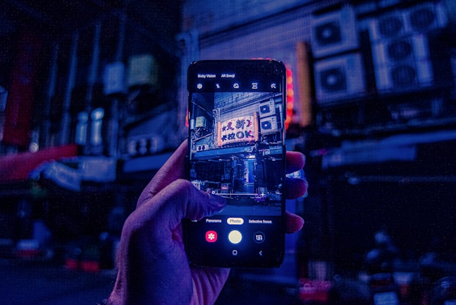 A hand holds a smartphone displaying a vibrant street scene captured in low light. Neon signs with apparent Japanese characters are visible in the photograph on the phone screen, while the background is dark and filled with indistinct urban elements such as air conditioning units and pipes.