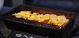 A black charcoal grill with a metal grate, cooking pieces of yellow-golden marinated chicken and sliced yellow vegetables, possibly squash or potatoes. The grill is emitting a warm glow from the fire underneath, which enhances the appearance of the food.