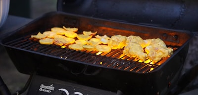 A black charcoal grill with a metal grate, cooking pieces of yellow-golden marinated chicken and sliced yellow vegetables, possibly squash or potatoes. The grill is emitting a warm glow from the fire underneath, which enhances the appearance of the food.