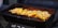A black charcoal grill with a metal grate, cooking pieces of yellow-golden marinated chicken and sliced yellow vegetables, possibly squash or potatoes. The grill is emitting a warm glow from the fire underneath, which enhances the appearance of the food.