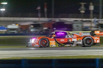 A high-performance race car speeds along a racetrack at night. The car is adorned with various sponsor logos and features a vibrant color scheme with prominent red and orange hues. Bright track lighting and blurred backgrounds enhance the sense of motion.