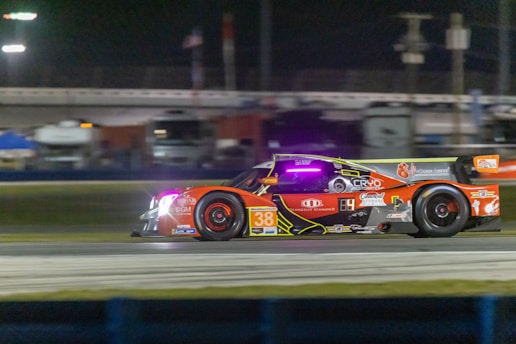 A high-performance race car speeds along a racetrack at night. The car is adorned with various sponsor logos and features a vibrant color scheme with prominent red and orange hues. Bright track lighting and blurred backgrounds enhance the sense of motion.