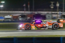 A high-performance race car speeds along a racetrack at night. The car is adorned with various sponsor logos and features a vibrant color scheme with prominent red and orange hues. Bright track lighting and blurred backgrounds enhance the sense of motion.