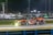 A sleek race car with a striking black and orange livery speeds along a well-lit track at night. The car is marked with the number 70 and features several sponsor logos including 'inception'. Its headlights are on, illuminating the track ahead, and a green rear spoiler adds contrast to its design. The background shows blurred lights and indistinct shapes, indicating motion.