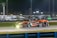 A sleek race car with a striking black and orange livery speeds along a well-lit track at night. The car is marked with the number 70 and features several sponsor logos including 'inception'. Its headlights are on, illuminating the track ahead, and a green rear spoiler adds contrast to its design. The background shows blurred lights and indistinct shapes, indicating motion.
