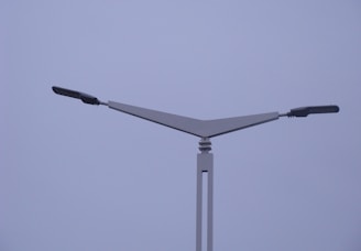 A modern street light with a minimalist design featuring two horizontal lamp arms extending outward in opposite directions from a triangular support structure. The background is a cloudy, overcast sky.