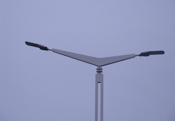 A modern street light with a minimalist design featuring two horizontal lamp arms extending outward in opposite directions from a triangular support structure. The background is a cloudy, overcast sky.