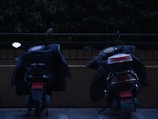 Two scooters are parked side by side, covered with what appears to be protective jackets draped over them. They are positioned in front of a railing with dense green foliage in the background. The image has a somewhat dim or shadowy quality, suggesting it might be taken in low light or twilight conditions.