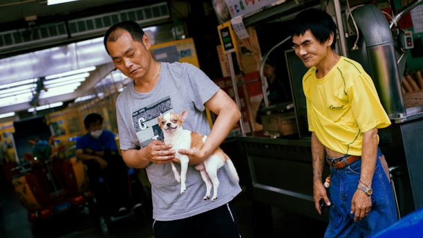 Two men are standing outside a shop. One man is holding a small dog, and the other is standing next to him, smiling. The attire of the men is casual. The background includes various shop signs and a glimpse of a market atmosphere.