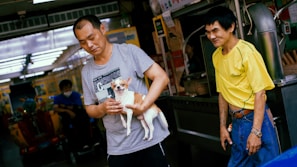 Two men are standing outside a shop. One man is holding a small dog, and the other is standing next to him, smiling. The attire of the men is casual. The background includes various shop signs and a glimpse of a market atmosphere.