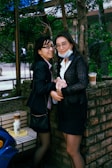 Two individuals are standing close to each other in an outdoor setting. Both are wearing business attire and glasses. One person is holding a beverage, while the other has a purse. They are positioned near a brick wall with trees and greenery in the background. There are two drinks placed on a nearby bench.