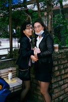 Two individuals are standing close to each other in an outdoor setting. Both are wearing business attire and glasses. One person is holding a beverage, while the other has a purse. They are positioned near a brick wall with trees and greenery in the background. There are two drinks placed on a nearby bench.