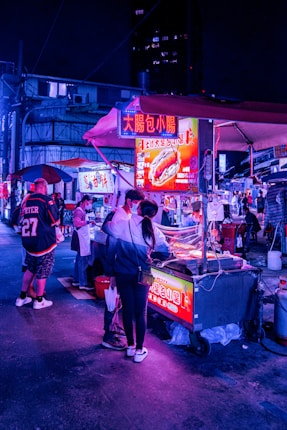 A bustling night market scene with vibrant neon lights illuminating food stalls. People are gathered around a street food vendor, examining the offerings. An assortment of signs and banners, written in Chinese, advertise various foods. The atmosphere is lively and colorful, with a mix of urban and cultural elements.