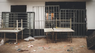 Metal framed structures are arranged against a wall with two tables in the foreground. The floor shows scattered wires and debris, suggesting a workshop or construction setting.