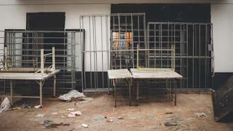 Metal framed structures are arranged against a wall with two tables in the foreground. The floor shows scattered wires and debris, suggesting a workshop or construction setting.