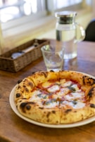A rustic, freshly baked pizza with a thick, bubbly crust sits on a wooden table. The toppings include melted cheese, green herbs, and a rich red tomato sauce. A clear glass and a pitcher of water are also on the table, along with a woven basket in the background containing utensils.