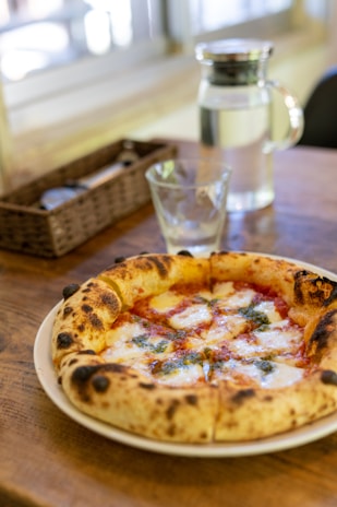 A rustic, freshly baked pizza with a thick, bubbly crust sits on a wooden table. The toppings include melted cheese, green herbs, and a rich red tomato sauce. A clear glass and a pitcher of water are also on the table, along with a woven basket in the background containing utensils.