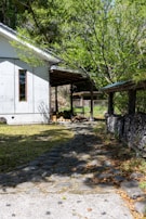 A serene yard featuring a stone pathway leading to a rustic structure partially covered by a roof. The area is framed by lush greenery with trees surrounding the property. A stack of firewood is neatly arranged under a wooden shelter along the right side. Sunlight filters through the leaves, casting dappled shadows on the ground.