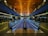 A spacious and modern airport terminal featuring a long moving walkway. The ceiling is wooden with lights embedded, creating a warm atmosphere. LED lights run along the sides of the walkway, with gates and seating areas visible on either side. Information displays and signs are illuminated in the distance.
