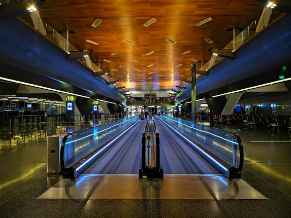 A spacious and modern airport terminal featuring a long moving walkway. The ceiling is wooden with lights embedded, creating a warm atmosphere. LED lights run along the sides of the walkway, with gates and seating areas visible on either side. Information displays and signs are illuminated in the distance.