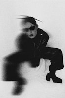 A person dressed in dark, avant-garde clothing is partially obscured by a blur effect. The person is wearing oversized sunglasses and has their hair styled upward. The background appears to be a plain, light-colored surface. The overall image quality is grainy, giving it a vintage or artistic feel.